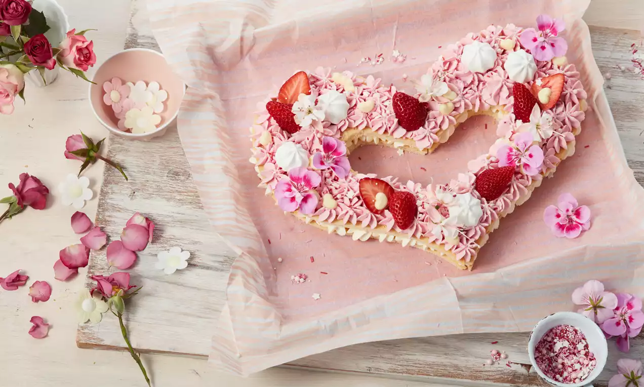 Herzförmige Cremetorte aus zwei Teigschichten mit rosa Sahnetupfen, frischen Erdbeeren, Baisers und Blüten auf rosa Papier drapiert