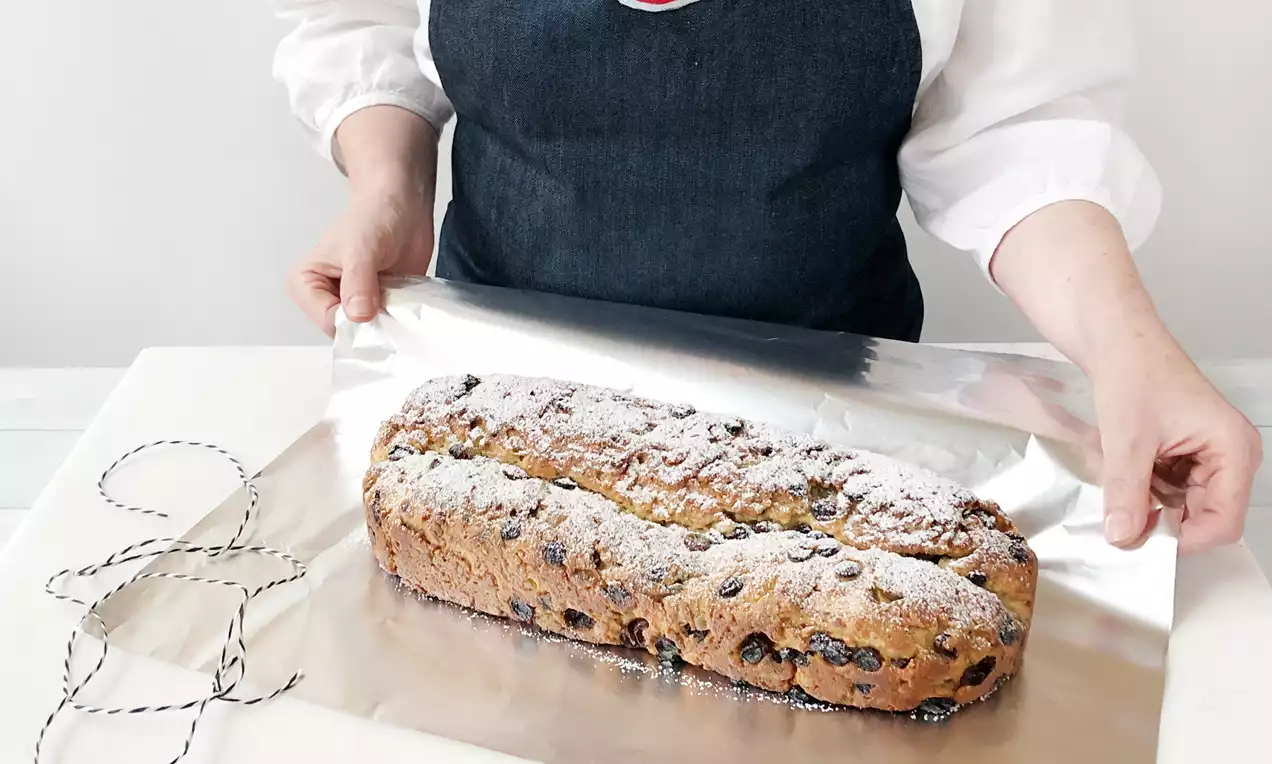 Goldbrauner Stollen mit Puderzucker bestäubt auf glänzender Aluminiumfolie, daneben weiß-schwarze Schnur, Hände in Schürze arrangieren