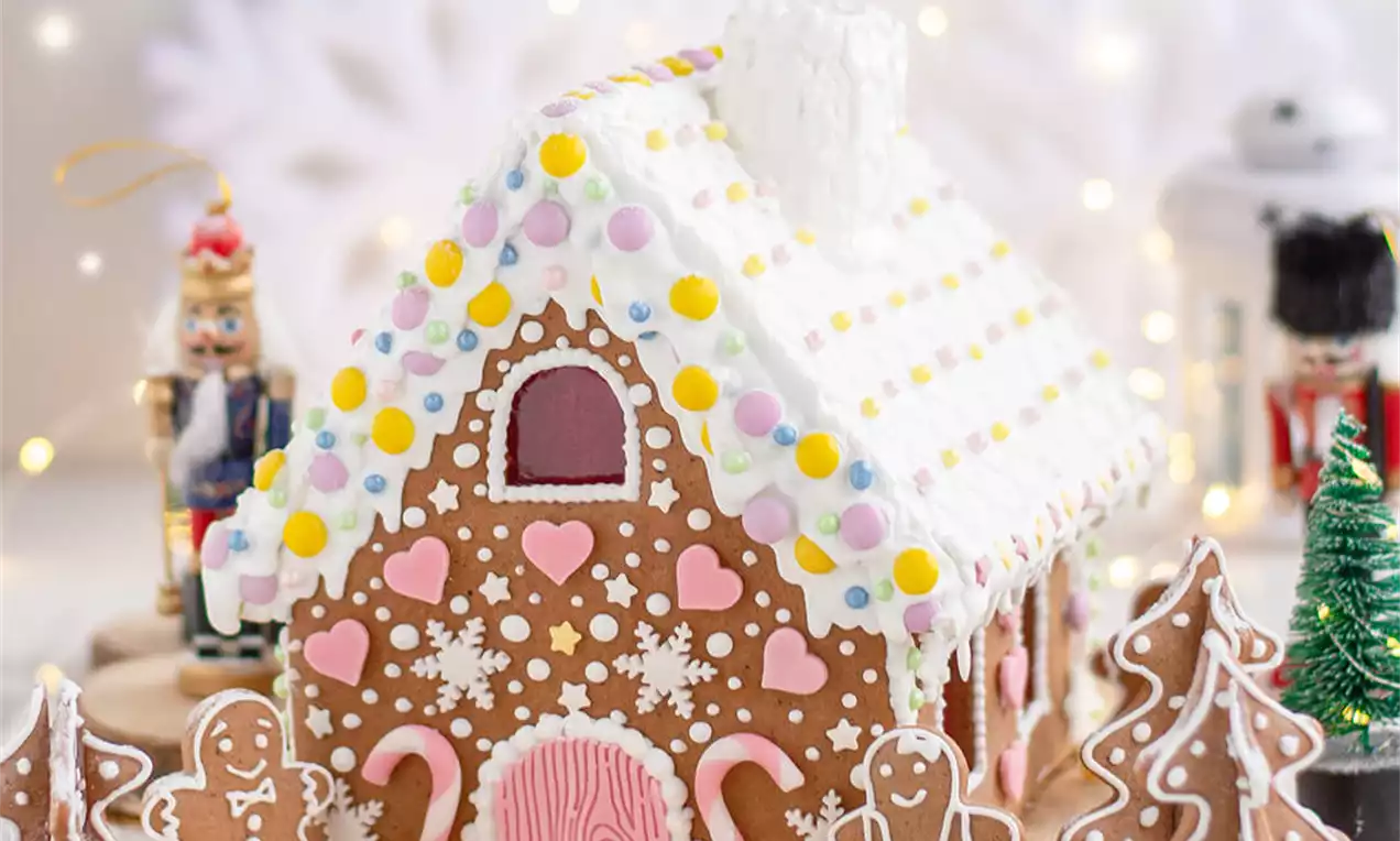 Medena kućica smeđe boje s bijelim detaljima, šarenim bombonima, ružičastim vratima i ukrasnim borovima na drvenoj podlozi okruženoj zimskom dekoracijom