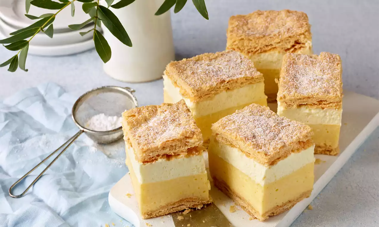 Quadratische Cremeschnitten mit goldbrauner Blätterteigkruste, luftig gelber Vanillecreme und weißer Sahneschicht auf weißer Platte