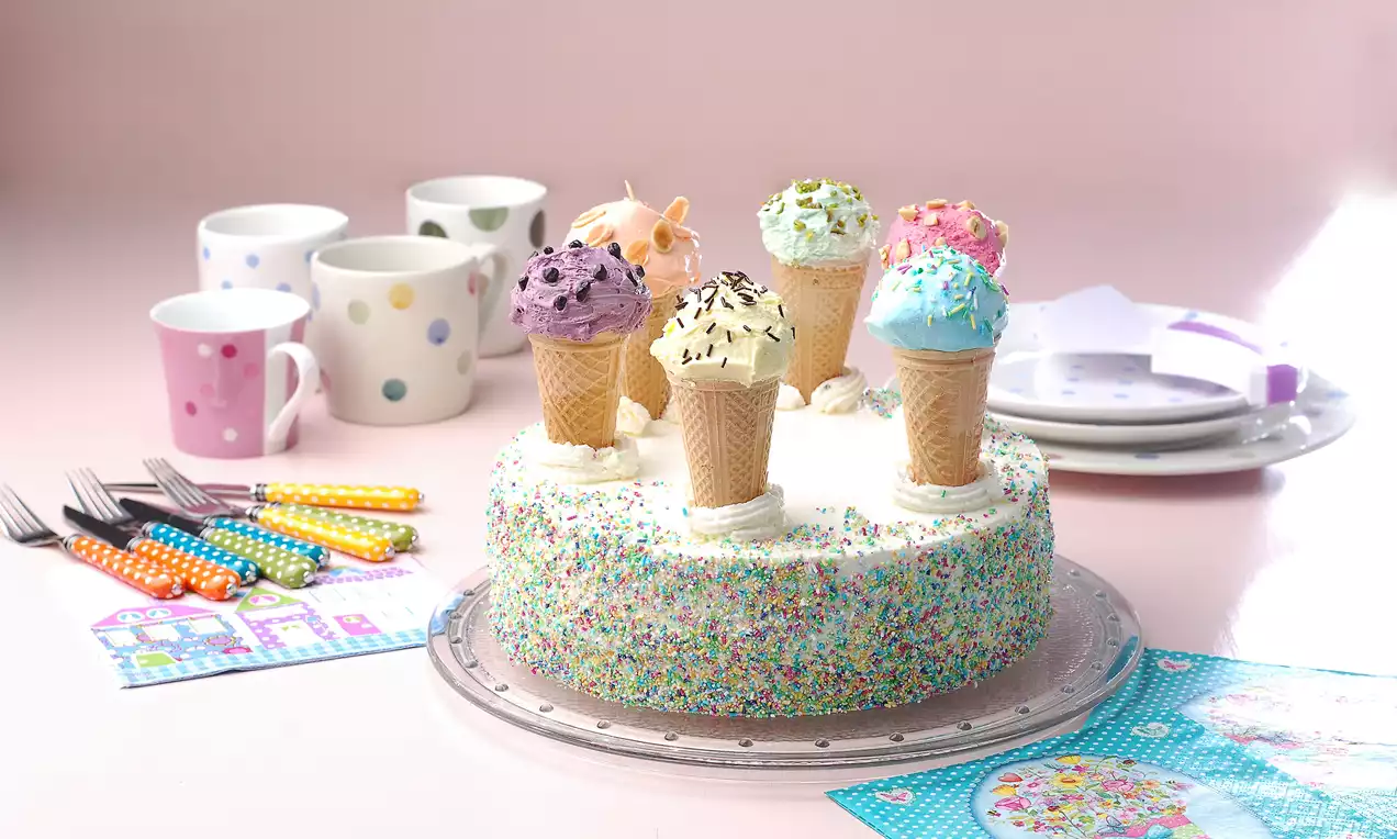 Yuvarlak beyaz pasta, krema kaplı kenarları renkli şekerlemelerle süslenmiş, üstünde külahlı dondurmalarla dekor edilmiş şekilde sunulmuş
