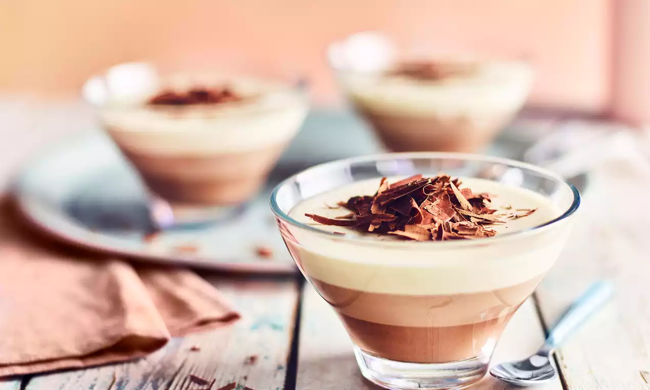 Layered dessert with smooth milk and white chocolate tiers in a glass bowl topped with thin, curled chocolate shavings on a rustic wooden table