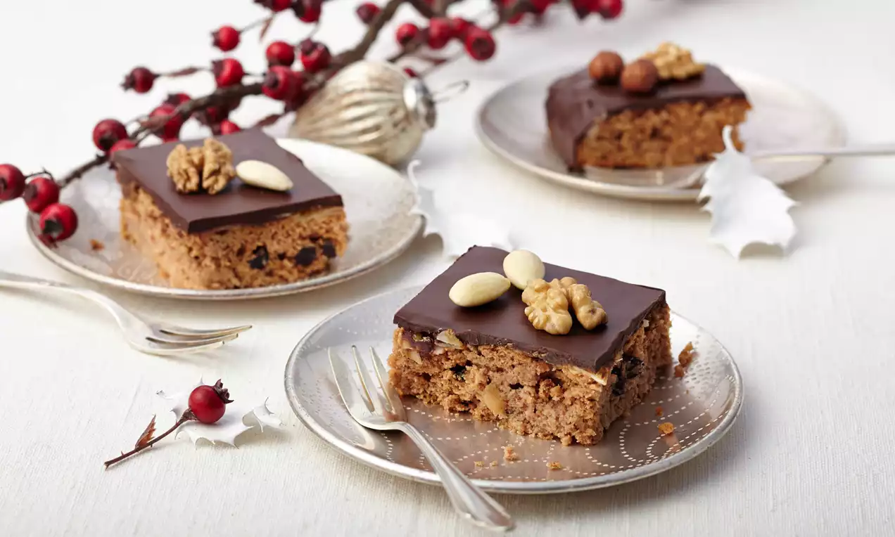 Honigkuchenstücke mit glänzender Schokoladenglasur, dekoriert mit Walnüssen und Mandeln auf silbernen Tellern, umgeben von roten Beerenzweigen