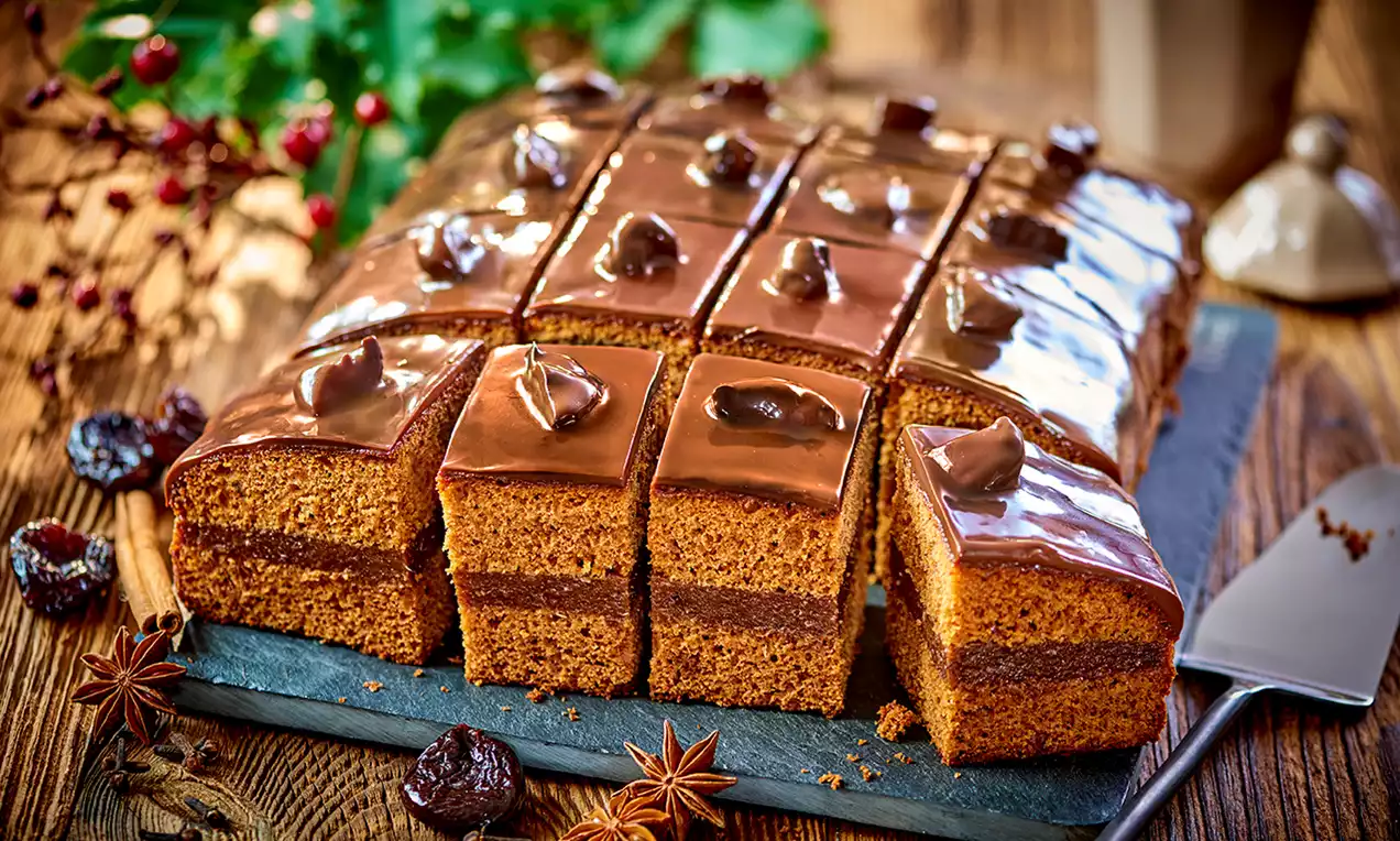 Kwadratowe kawałki śliwkowego piernika z daktylami, pokryte lśniącą czekoladą, ozdobione śliwkami, ułożone na ciemnej desce