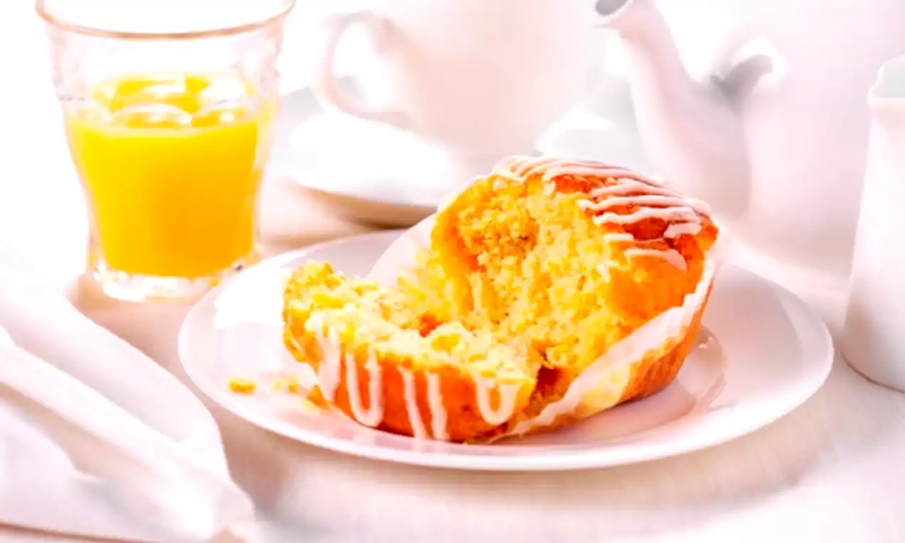 A golden orange and carrot muffin with a soft crumbly texture and white icing drizzles, served on a white plate with tea and juice