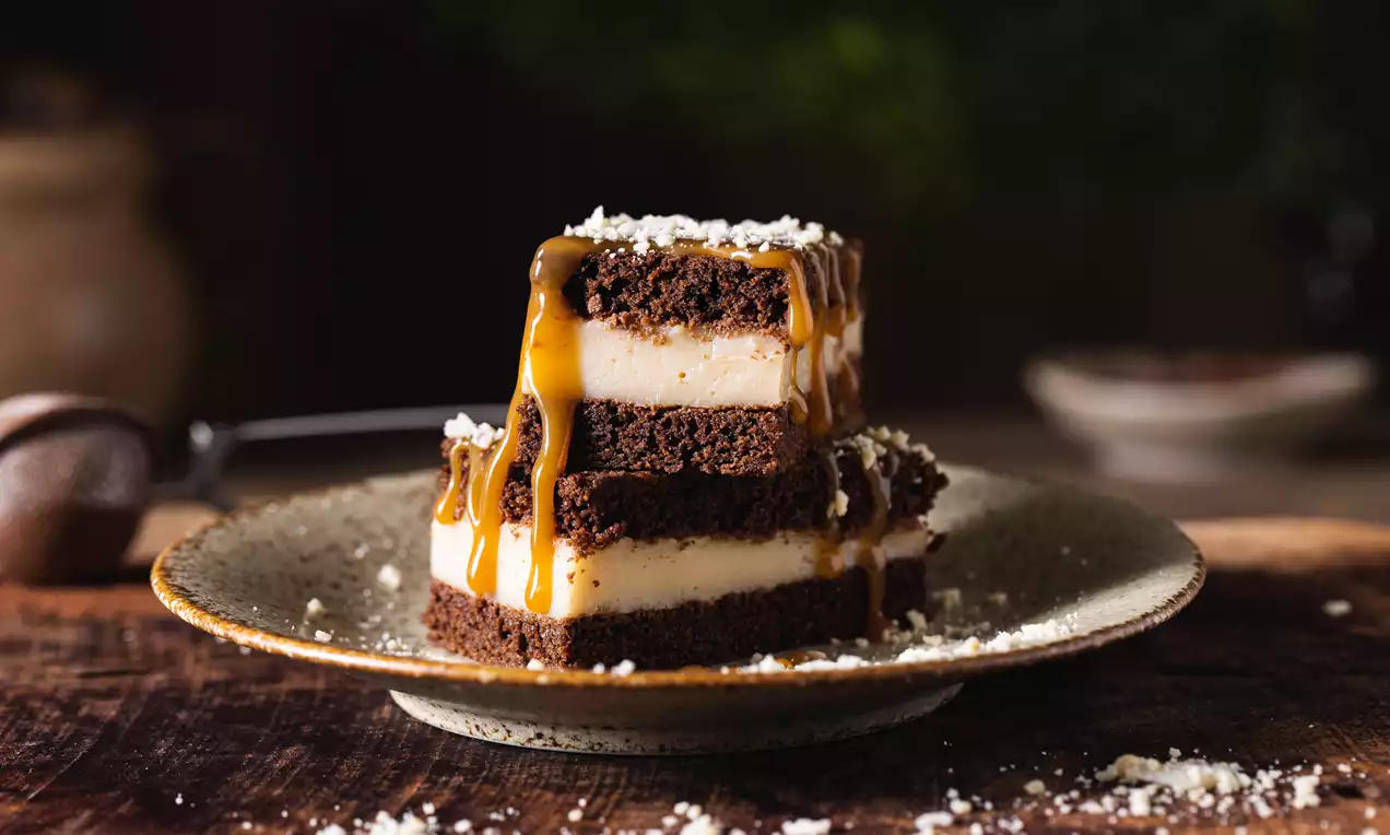 Tmavohnedé brownie vrstvy oddelené svetlým mandľovým pudingom, poliatu karamelom a posypanú drvenými mandľami na keramickom tanieri