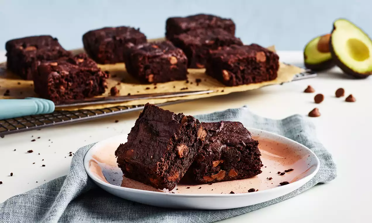 Tumma, kostea brownie, jossa kaakaon sävyistä marmorintäplää, avokadon vieressä vaaleanpunaisella lautasella sinisen liinan päällä