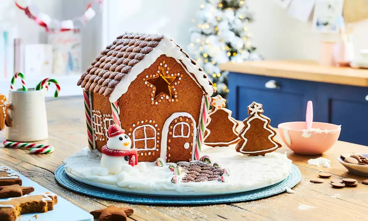 Brown gingerbread house with white icing details, candy cane pillars, chocolate roof tiles, snowy base, and festive tree decor