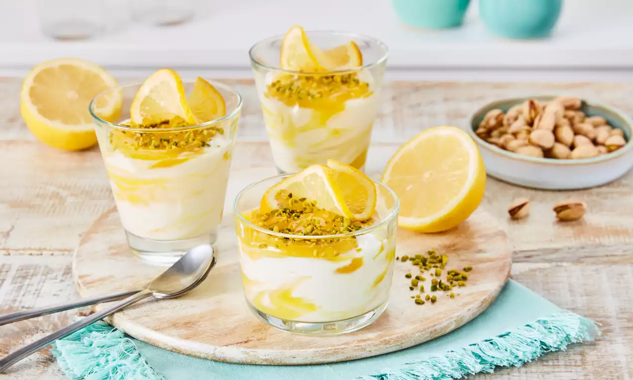 Gläser mit cremigem Zitronenquark, gelben Zitronenstreifen, Pistazienstückchen und Zitronenscheiben auf einem Holzbrett mit türkisfarbener Stoffserviette