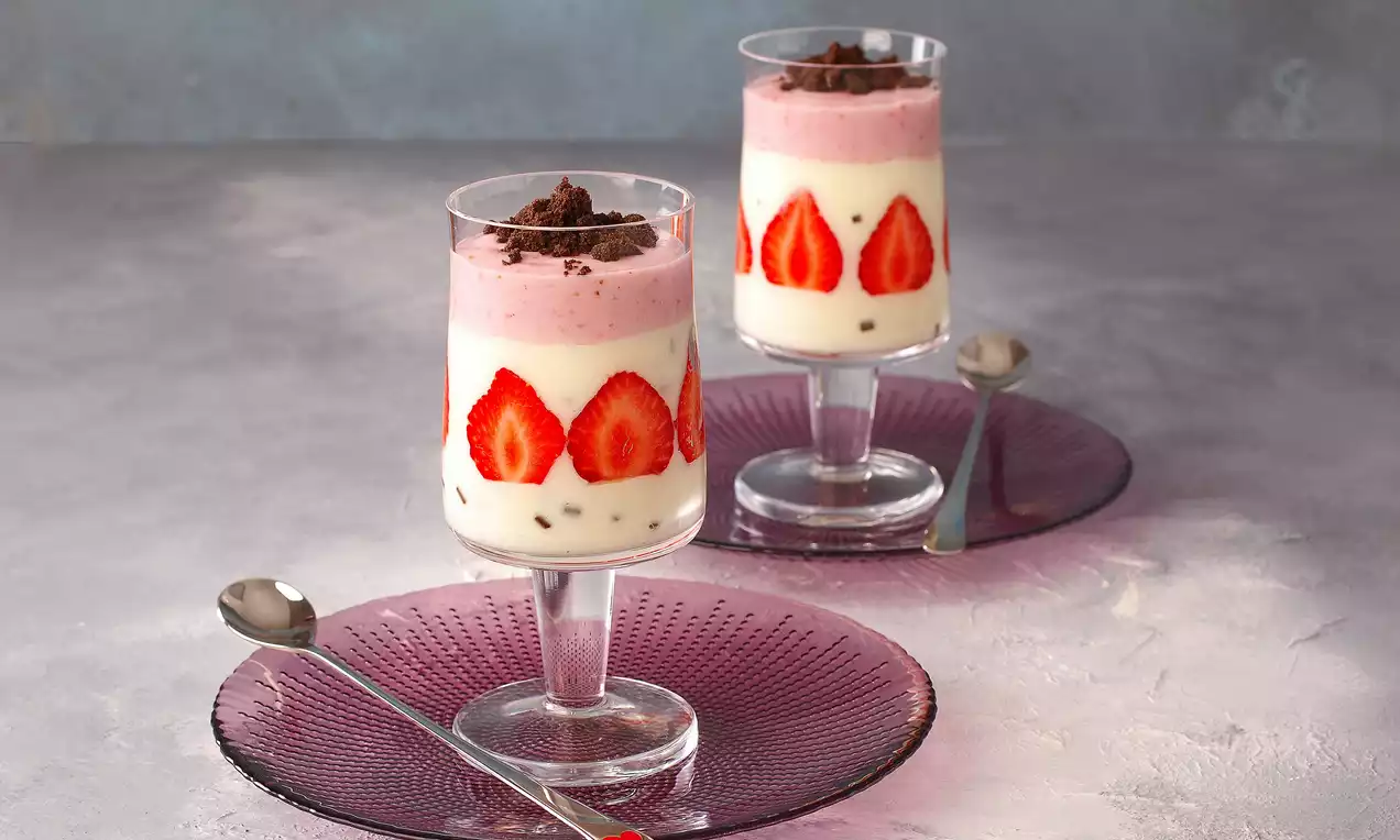 Kat kat dizilmiş çilek dilimleri, açık pembe ve beyaz tatlı kremayla dolu cam kup, üstünde ufalanmış çikolata parçalarıyla sunulmuş