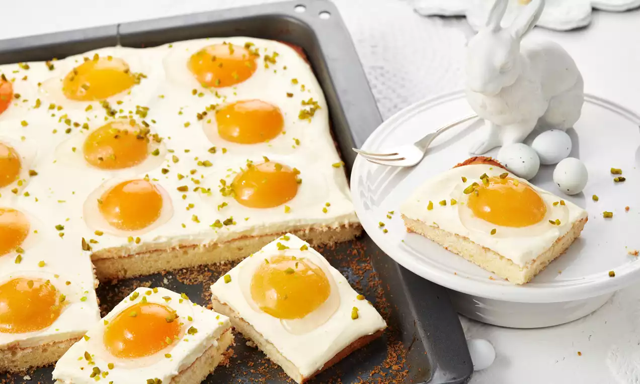 Spiegeleikuchen mit goldbraunem Biskuitboden, weiß-gelber Cremefüllung und ovalen, dottergelben Zuckereiern auf hellblauem Teller