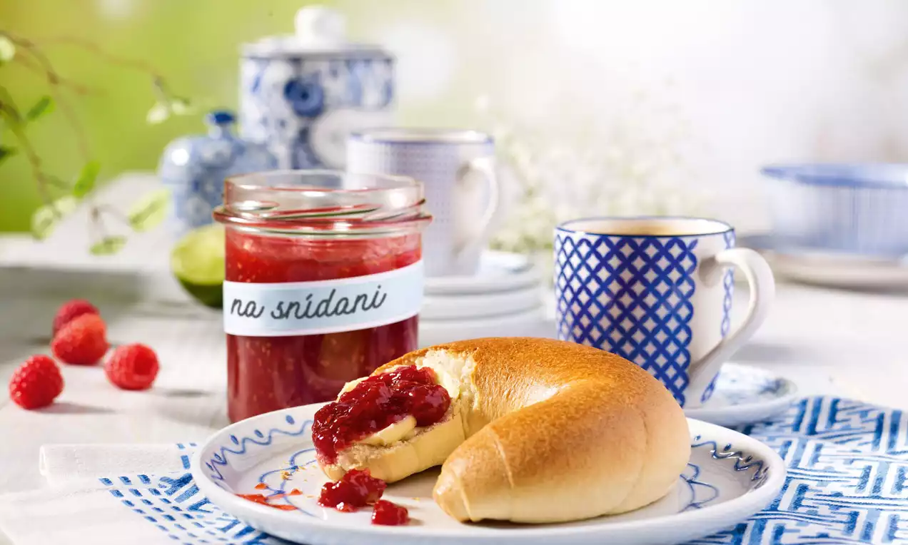 Sklenice malinovo-meruňkového džemu zářivě červené barvy vedle zlatavého loupáku na talíři s modrobílým vzorem v prosvětleném prostředí