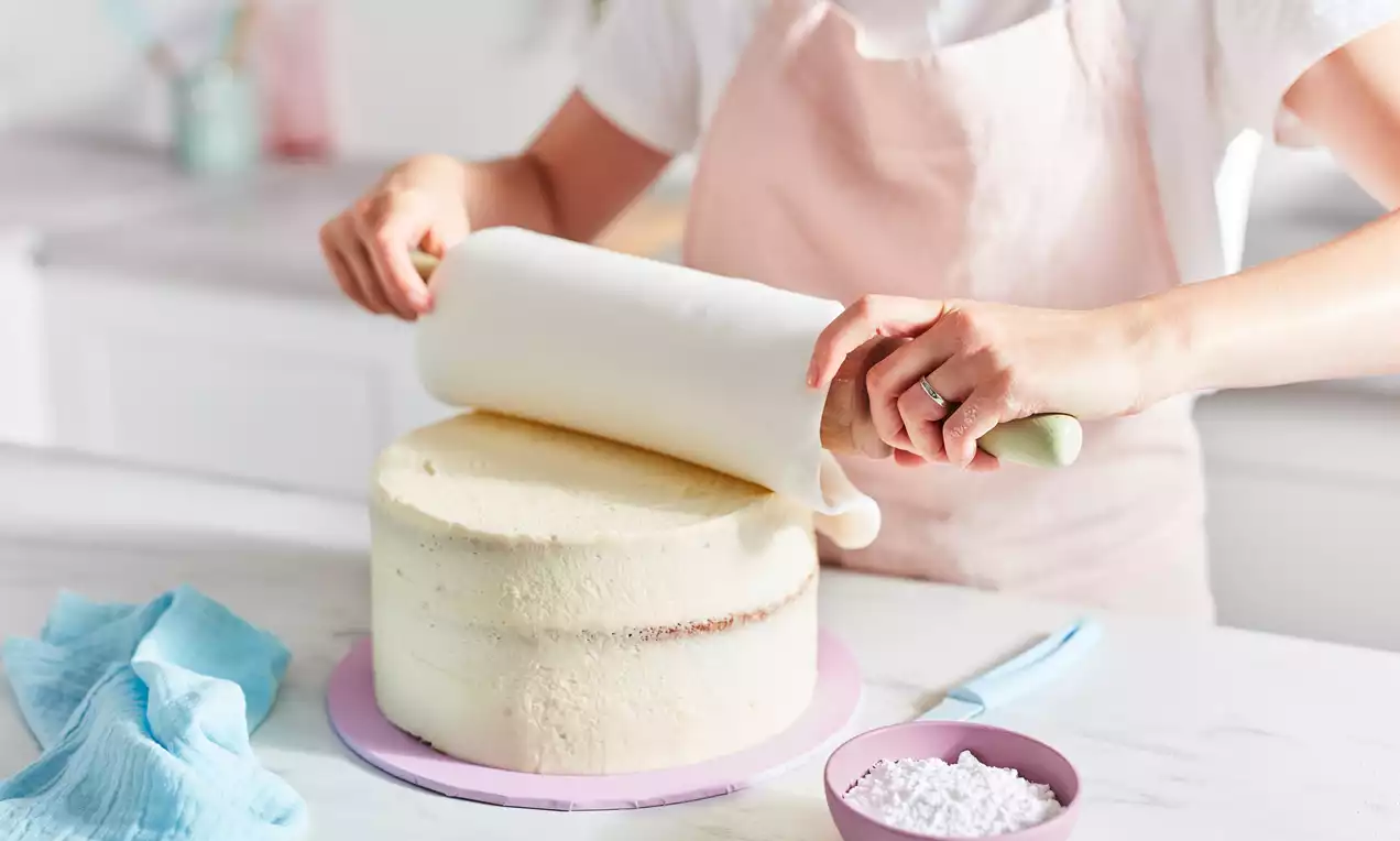 Smooth white fondant being rolled over a tall round cake with creamy beige frosting on a pastel purple board in a bright kitchen