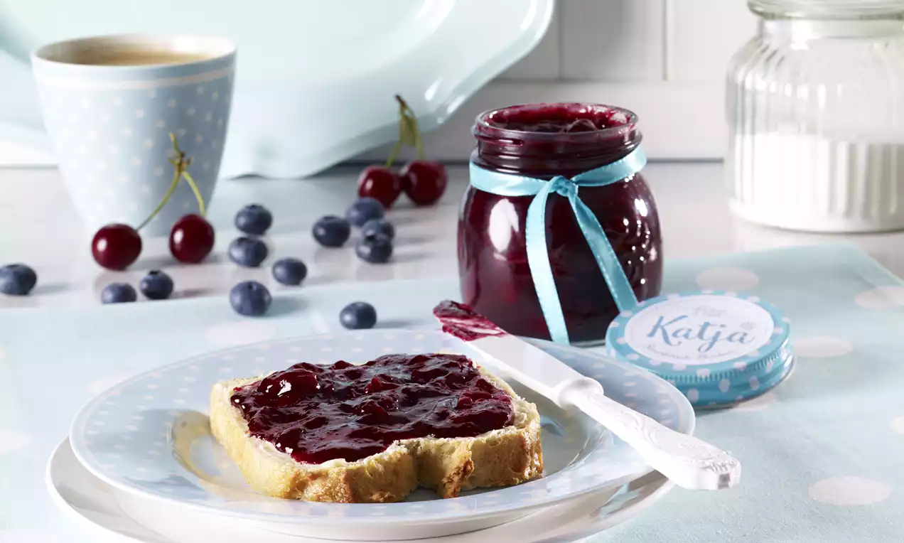 Conserva rosso intenso in vasetto di vetro con fiocco azzurro, spalmata su una fetta di pane irregolare accanto a coltello e frutti