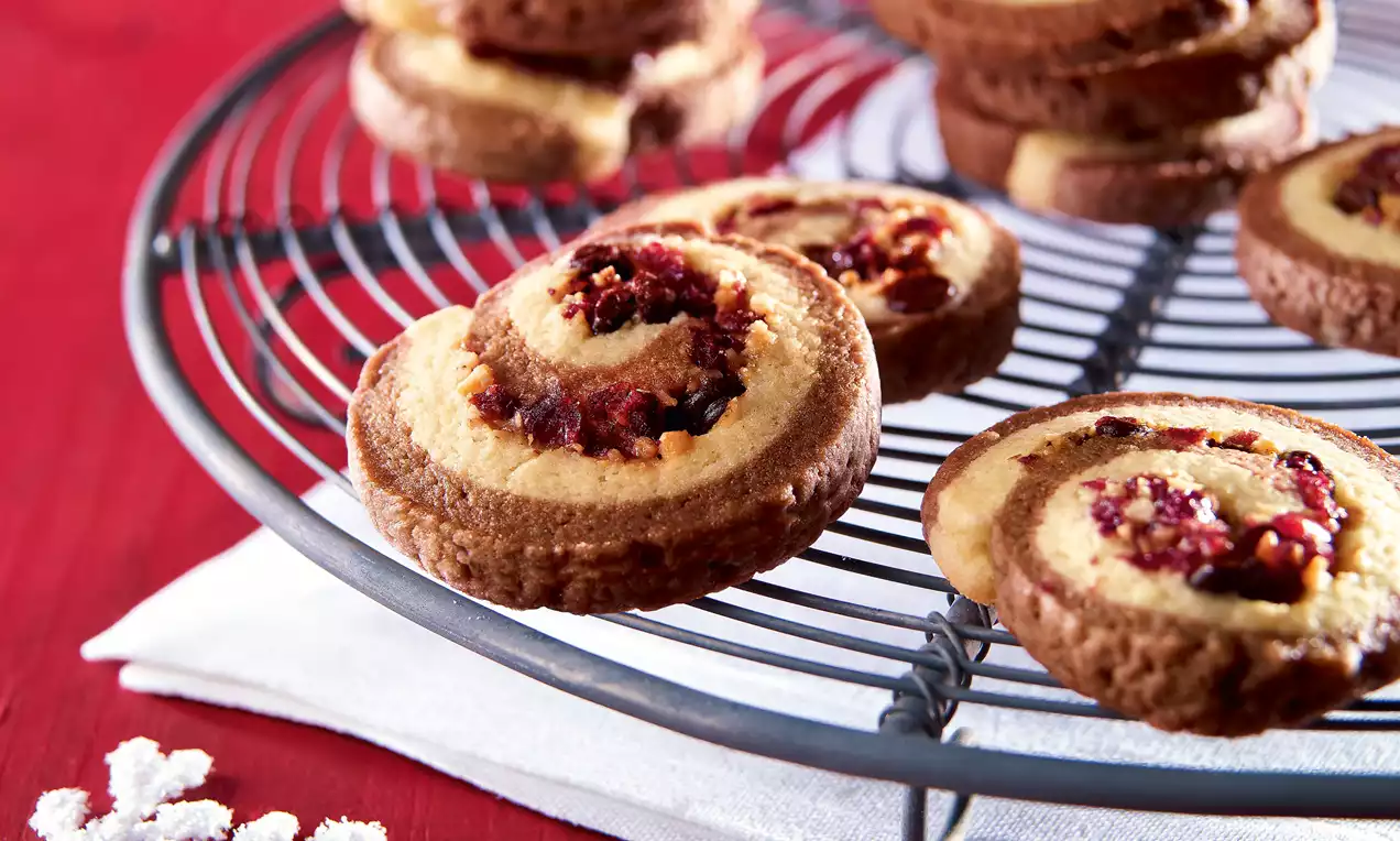 Cranberry-Schoko-Taler mit spiraliger Struktur aus hellem Mürbeteig, braunem Schokoladenrand und roten Cranberry-Stückchen auf einem Gitter