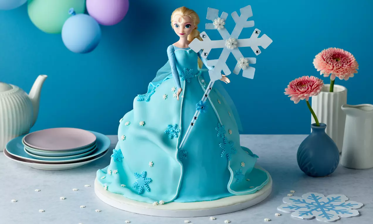 Torte in Form einer blau glänzenden Prinzessinnenrobe, dekoriert mit Schneeflocken, auf weißem Teller neben zarten Blumen und Geschirr