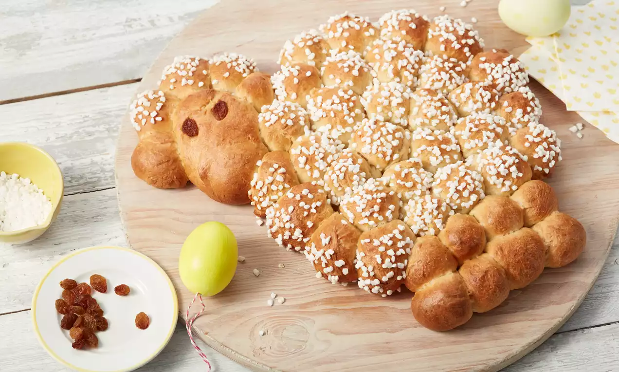 Osterlamm aus goldbraunem Hefeteig mit Hagelzucker dekoriert, auf einem runden Holzbrett neben gelbem Ei, Rosinen und Mehl