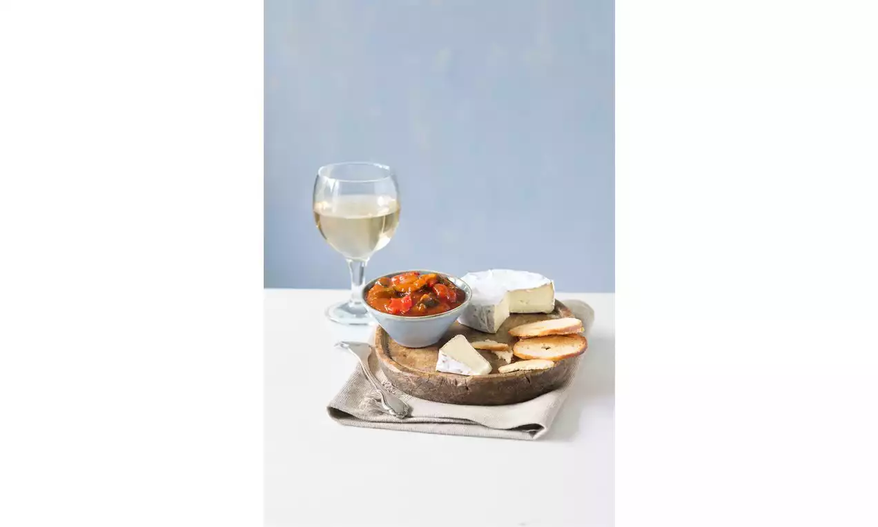 Zeleninový reliš v šedom miske s červenými a oranžovými tónmi, vedľa syrov a opečeného chleba na drevenom podnose s pohárom bieleho vína