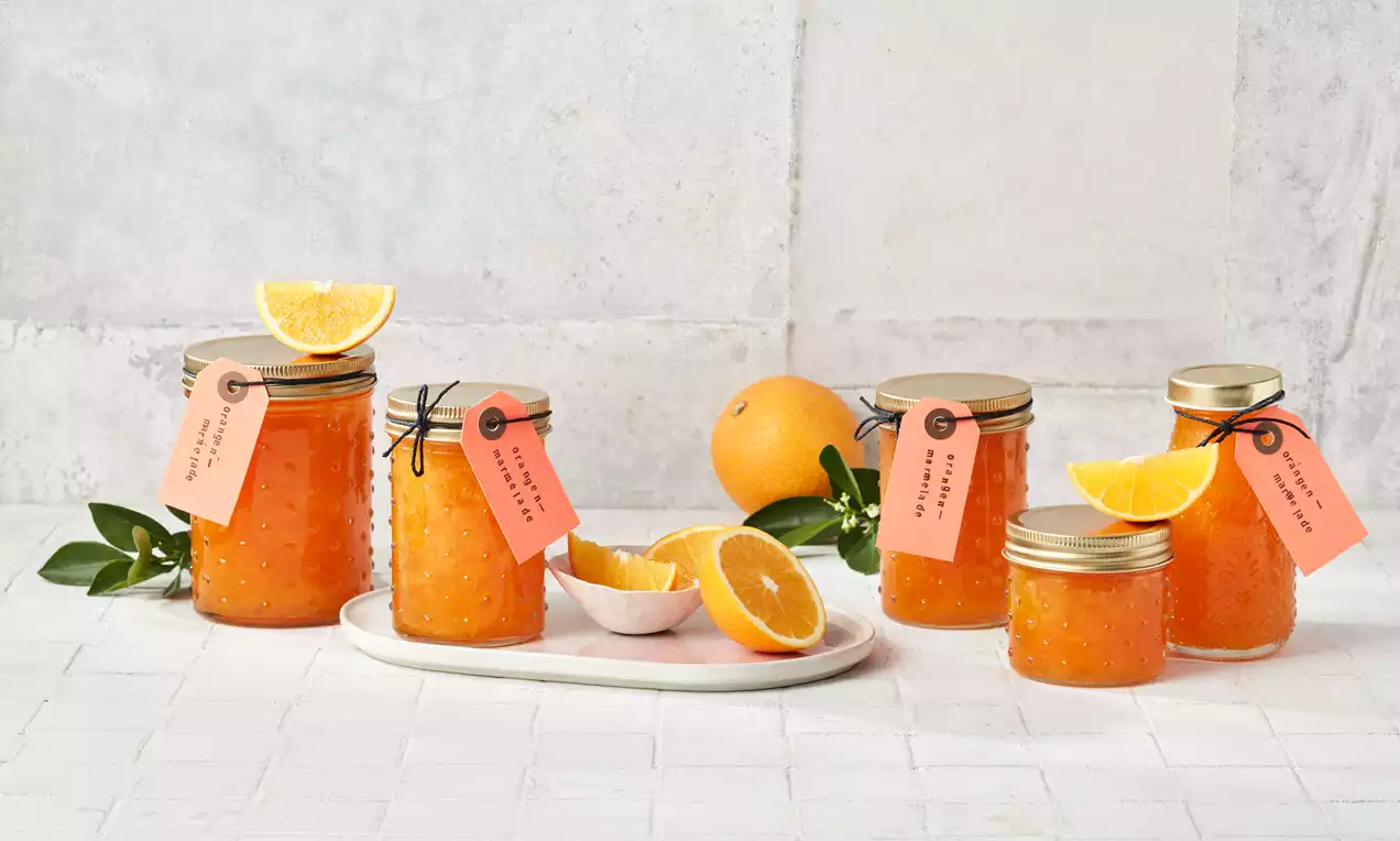 Gläser mit goldorangefarbener Orangenmarmelade und goldenen Deckeln, verziert mit rosafarbenen Anhängern, umgeben von Orangenhälften und Blättern