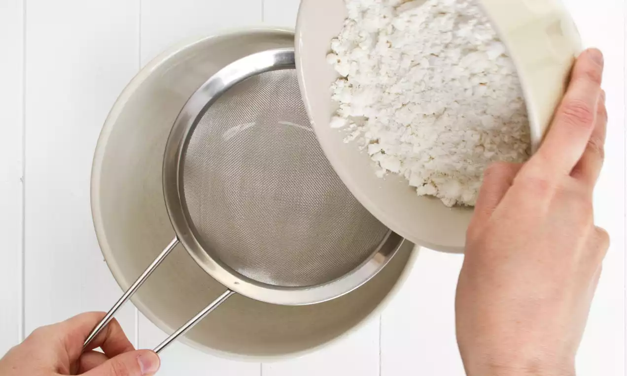 White flour falling from a cream bowl into a fine metal sieve held over a beige mixing bowl on a white wooden surface
