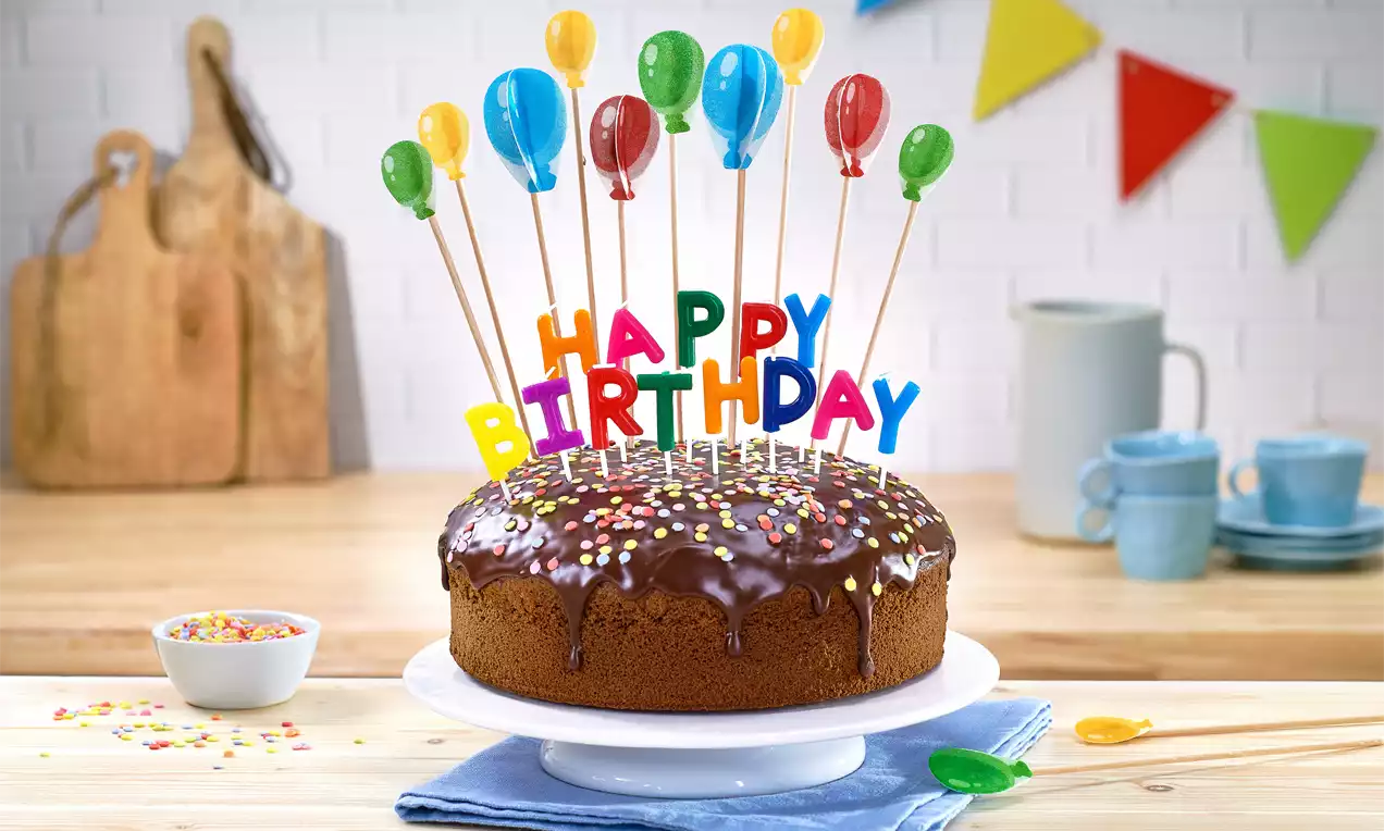 Schokoladenkuchen mit glänzendem Guss, bunten Streuseln und Happy-Birthday-Steckdeko mit Ballonmotiven auf weißer Tortenplatte