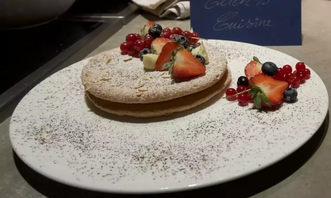 Rond amandelgebak bestrooid met poedersuiker, gegarneerd met aardbeien, rode bessen, blauwe bessen en witte chocolade op een licht gespikkeld bord
