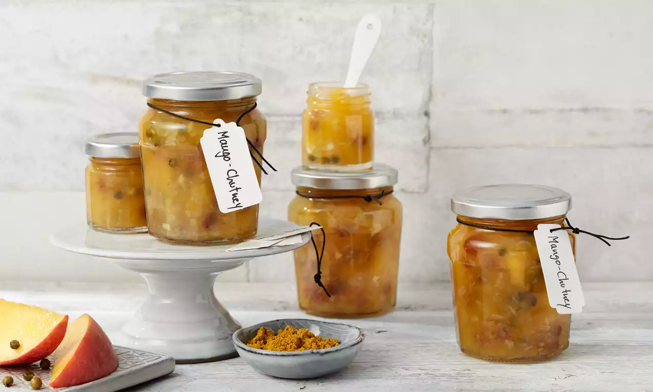 Staklenke različitih veličina s mango chutneyjem zlatnožute boje, uz male komadiće povrća, na rustikalnom bijelom stolu s tanjurom začina