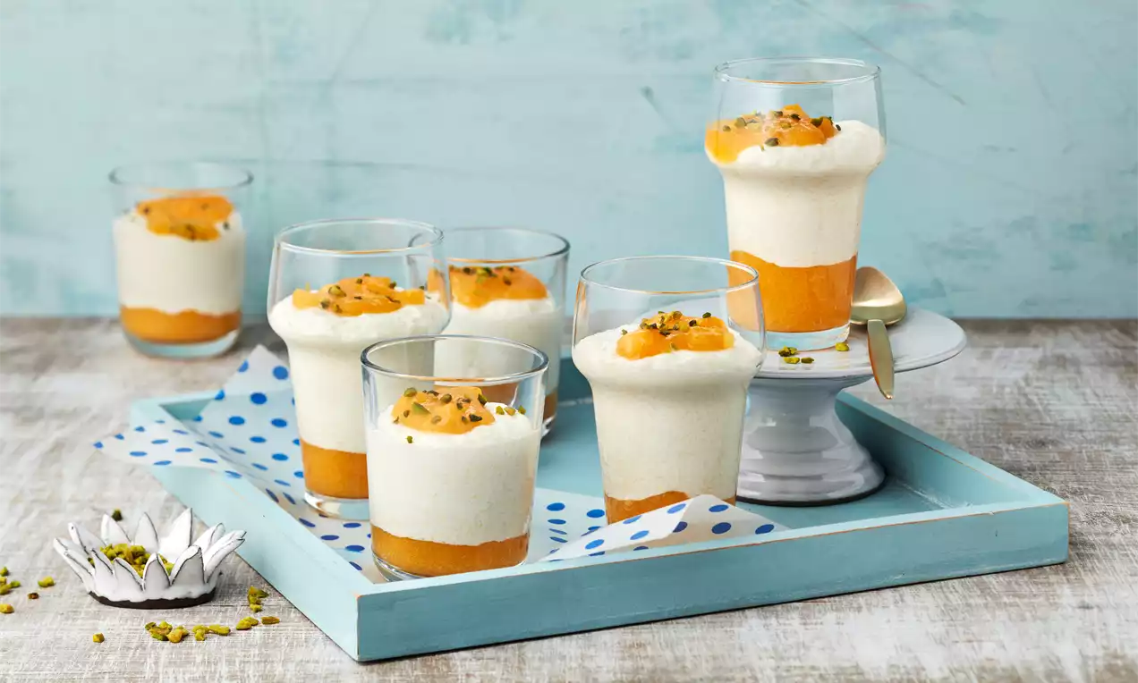 Gläser mit cremigem Grießbrei auf leuchtend orangefarbenem Pfirsichkompott, garniert mit Pistazienstücken auf einem hellblauen Tablett