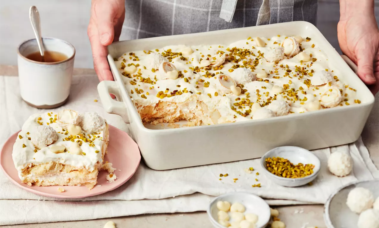 Raffaello-Tiramisu in heller Auflaufform mit Schichten aus Creme, Biskuit, weißen Pralinen und gehackten Pistazien auf beigem Tuch angerichtet
