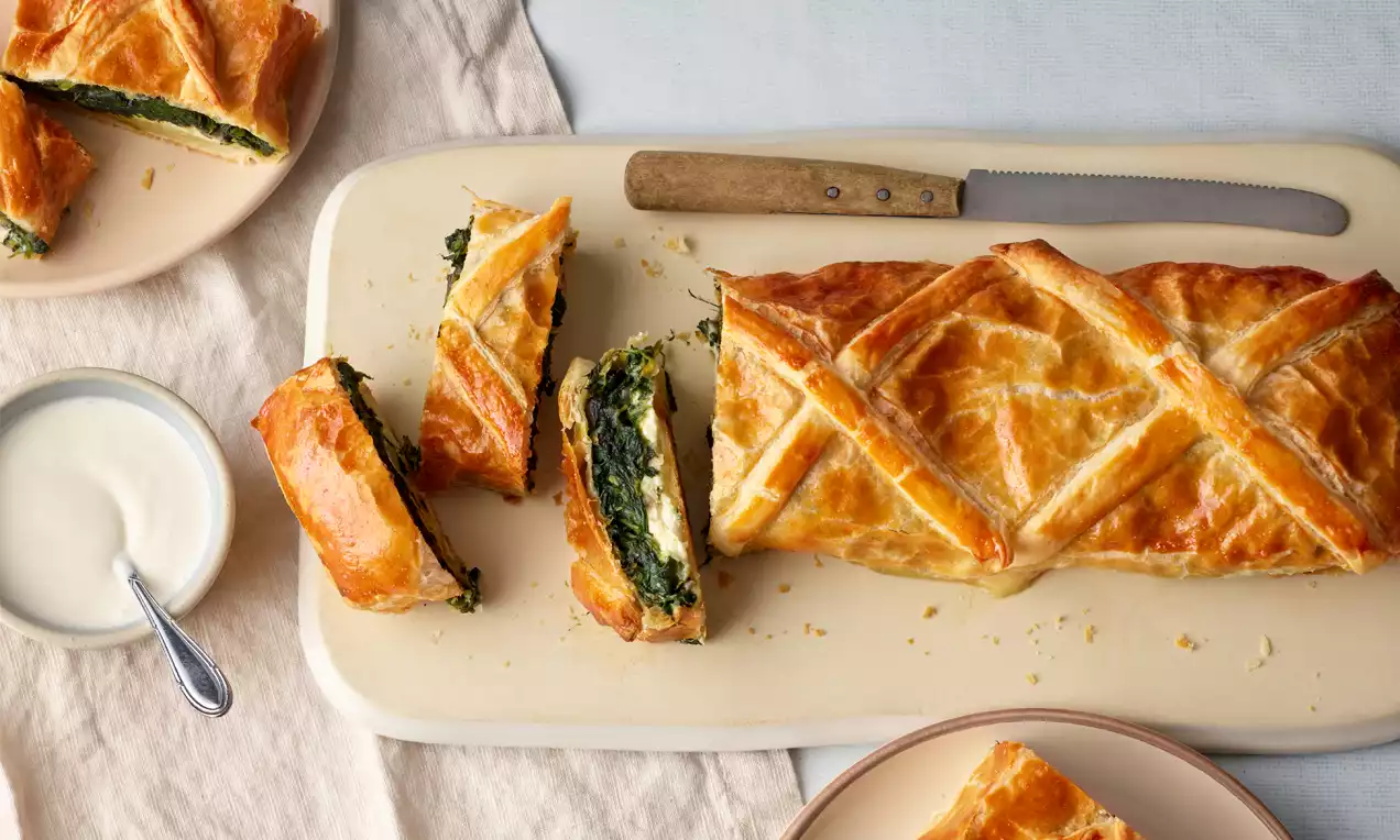 Goldbrauner Strudel mit filigranem Muster, gefüllt mit grüner Spinat-Schafskäse-Mischung, angerichtet auf einem rechteckigen Teller mit Messer