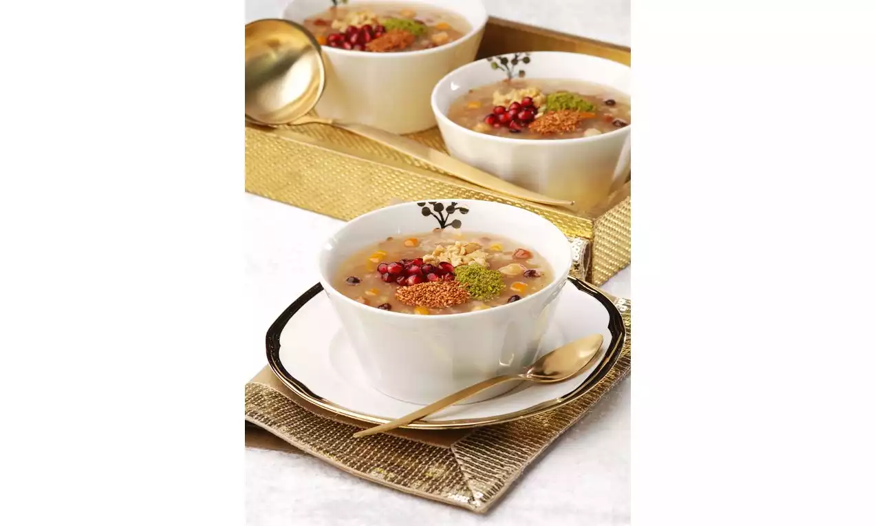 Beyaz porselen kasede sunulan aşure, nar taneleri, yeşil toz fıstık ve cevizle süslenmiş, metalik detaylı bir tabakta yer alıyor