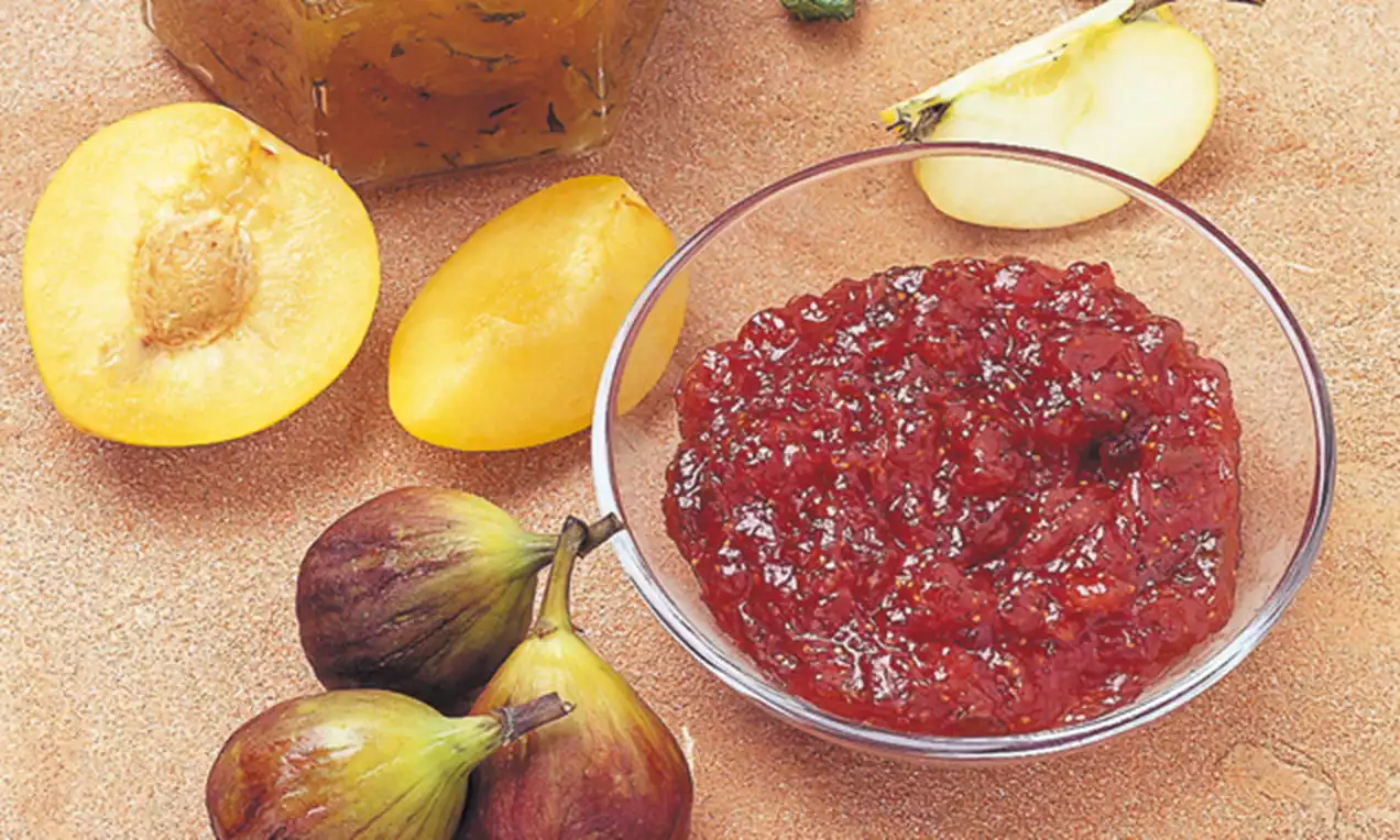 Composta rosso brillante in una ciotola trasparente accanto a fichi viola scuro, prugne gialle succose e un quarto di mela chiara