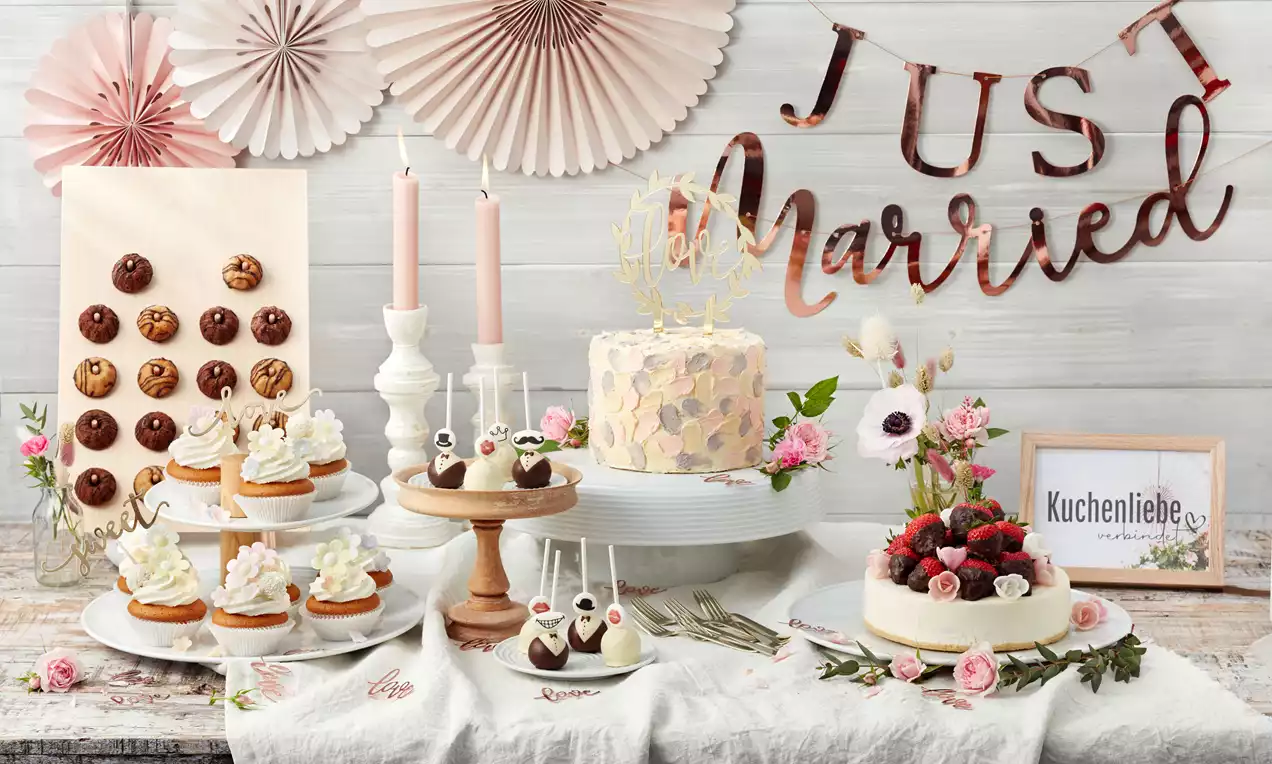 Eine Hochzeitstafel mit süßen Gebäcken, Kuchen und Torten auf Glas- und Keramikplatten, umgeben von rosa Blüten und Dekorationen in Pastelltönen
