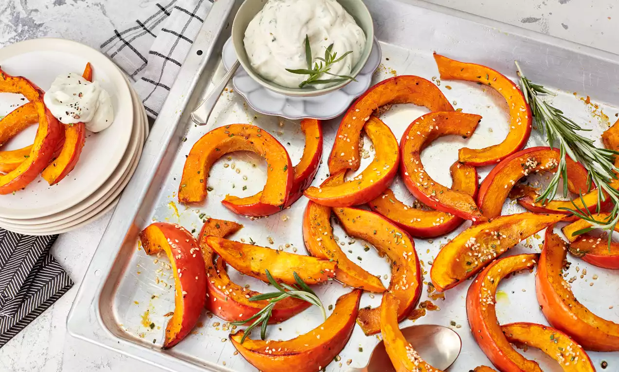 Geröstete orange Kürbisspalten mit goldbraunem Rand auf einem Backblech, garniert mit Kräutern, neben cremigem Dip in einer Schale