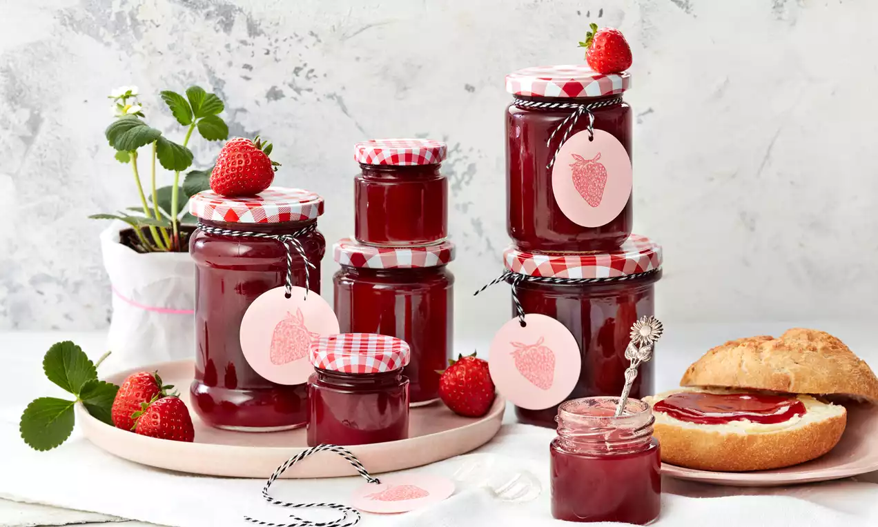 Erdbeerrotes Gelee in Gläsern mit karierten Deckeln und Etiketten, arrangiert neben frischen Erdbeeren und einem Brötchen mit Gelee
