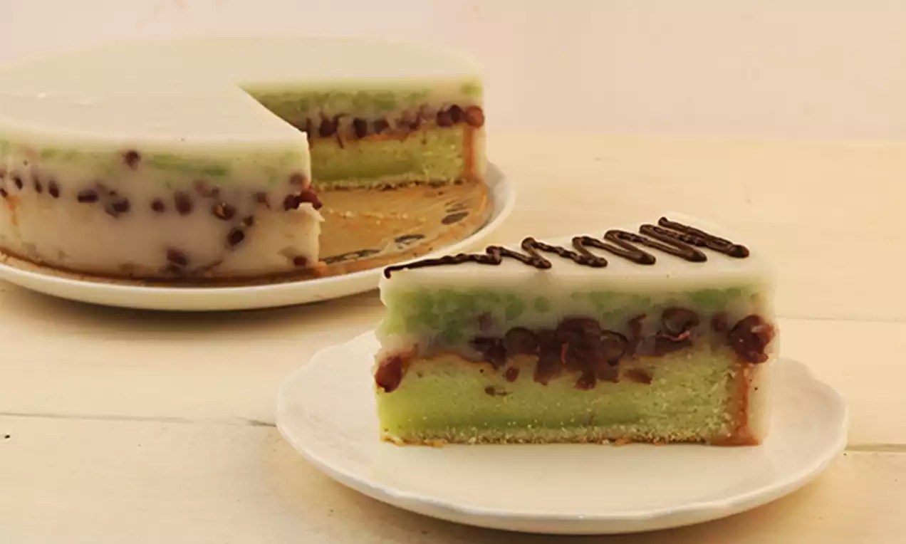 Creamy pandan agar cake with layers of translucent white jelly, green pandan custard, red beans, and a biscuit base on a white plate