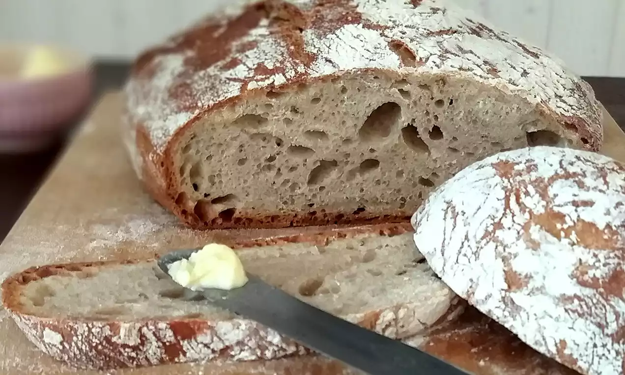 Kváskový chlieb s tmavozlatou chrumkavou kôrkou, posypaný múkou, s mäkkým pórovitým vnútrom, položený na doske vedľa noža s maslom