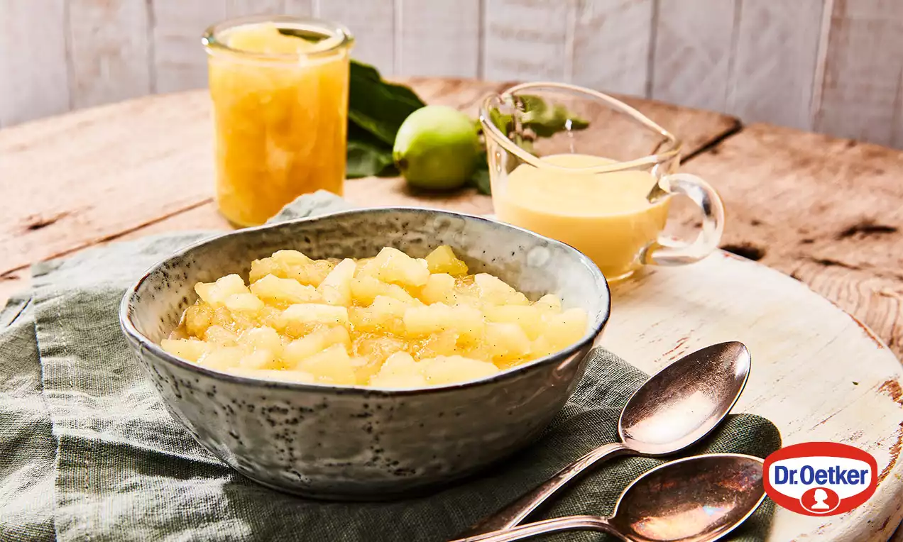 Svetložltý jablkový kompót s jemne lesklými kúskami v rustikálnej keramickej miske na zelenom obrúsku, obklopený lyžičkami a prílohami