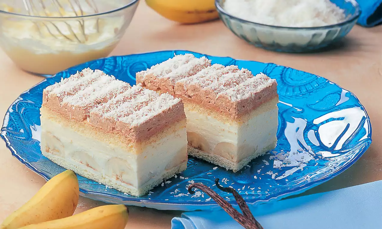 Zwei rechteckige Bananen-Kokosschnitten mit heller Creme, Kokosraspeln und goldbrauner Schicht auf blauem Teller neben gelben Bananen