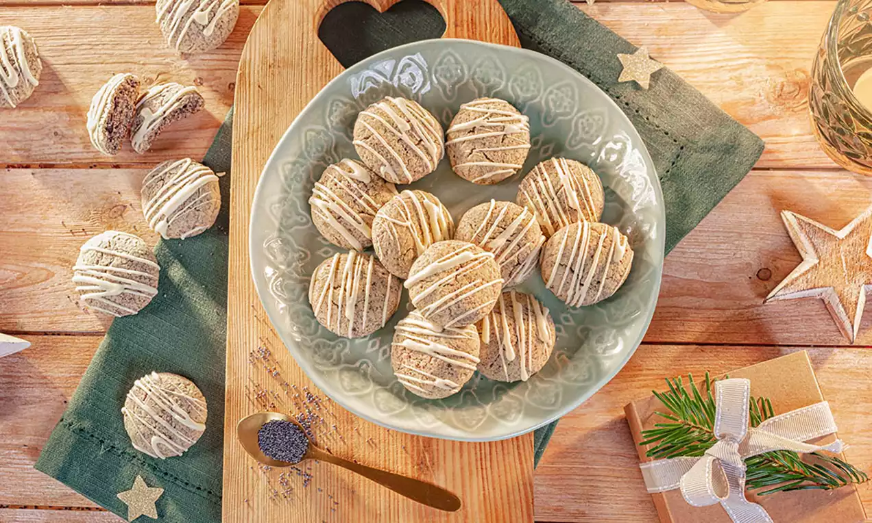 Runde, helle Mohn-Plätzchen mit feiner heller Glasur auf grünem Teller, arrangiert auf Holzbrett mit Deko aus Sternen und Geschenk