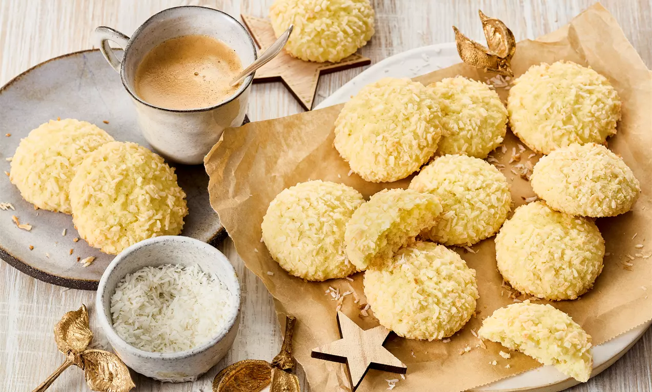 Gelbgoldene Kokos-Cookies mit flacher, leicht runder Form und grober Kokostextur auf braunem Backpapier mit Deko aus Holz und Kokosraspeln