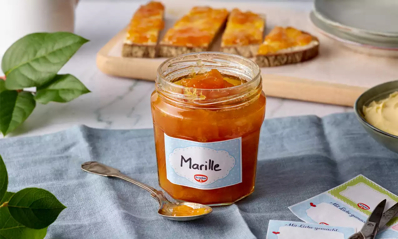 Glas mit leuchtend orangefarbener Marillenmarmelade, ein silberner Löffel, auf hellblauem Stoff neben brotig bestrichenen Scheiben