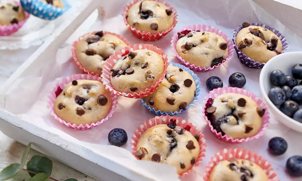 Kleine vegane Blaubeer-Muffins mit goldbrauner Oberfläche und dunklen Blaubeeren in bunten Papierförmchen auf weißem Tablett mit Blaubeeren