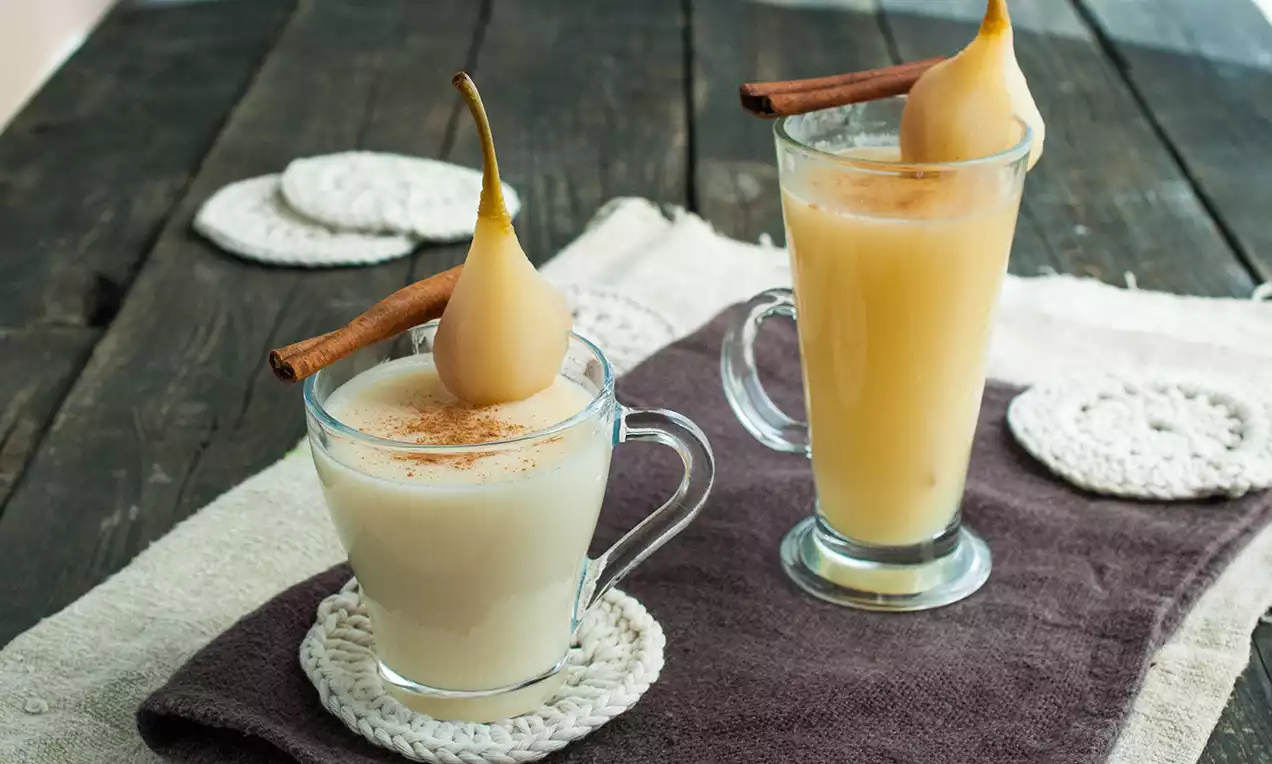 Krémový vanilkový nápoj světlé barvy v sklenici s uchy, zdobený celou vařenou hruškou a skořicovou tyčinkou na dřevěném stole