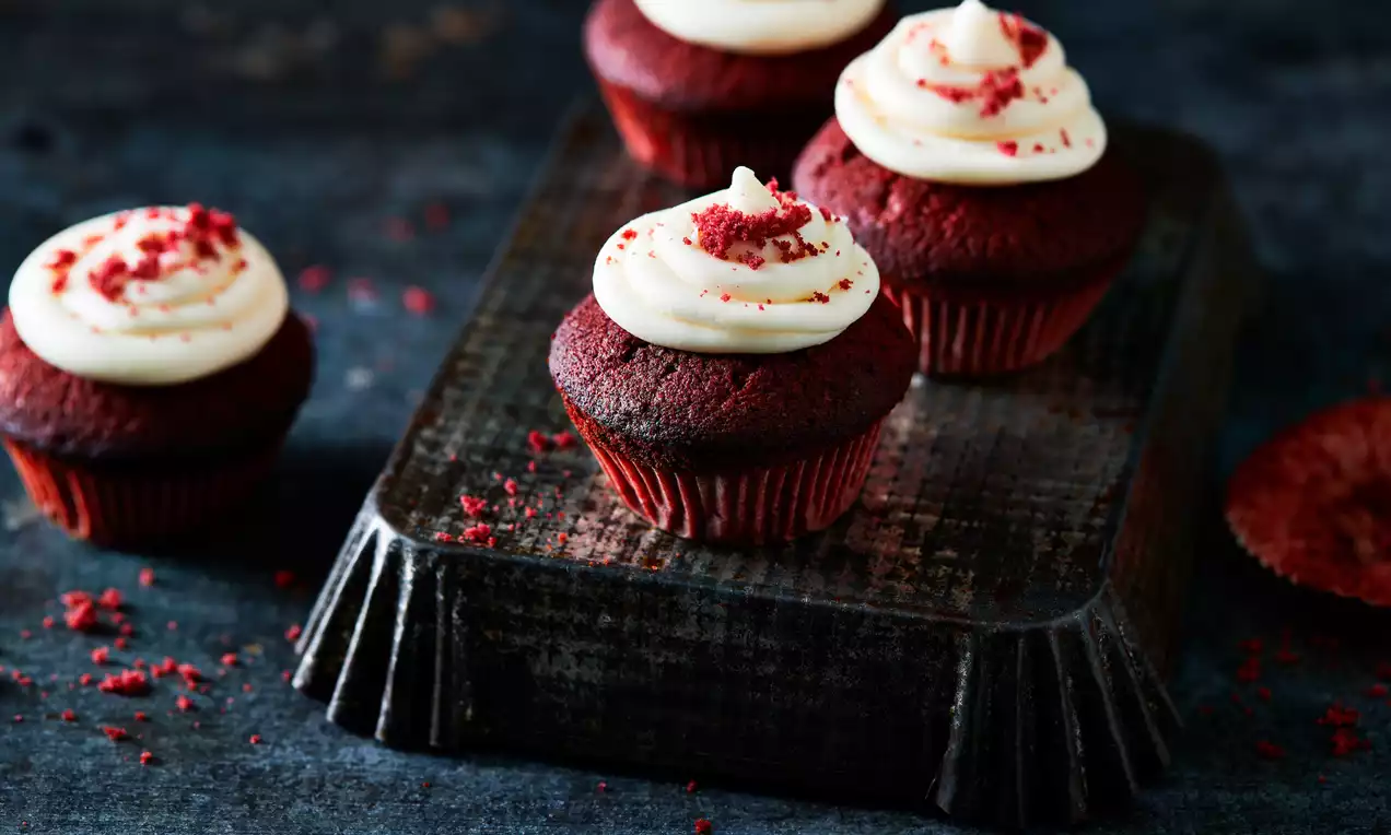 Cupcakes Red Velvet aux teintes rouge foncé garnis de crème blanche onctueuse et éclats rouge vif sur un plateau sombre rectangulaire