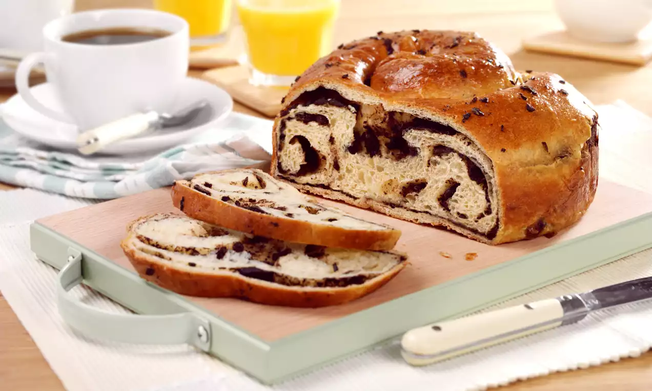 Golden, glossy chocolate swirl bread with dark cocoa ribbons inside, sliced on a wooden tray beside coffee, juice, and cutlery