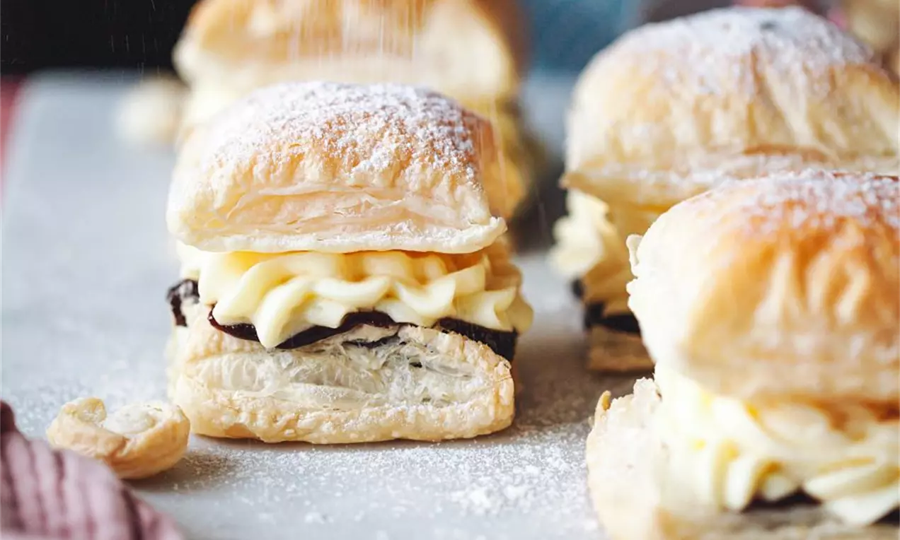 Nadýchané kocky zlatistého lístkového cesta plnené krémovou bledou plnkou a vrstvou tmavej čokolády, jemne posypané práškovým cukrom