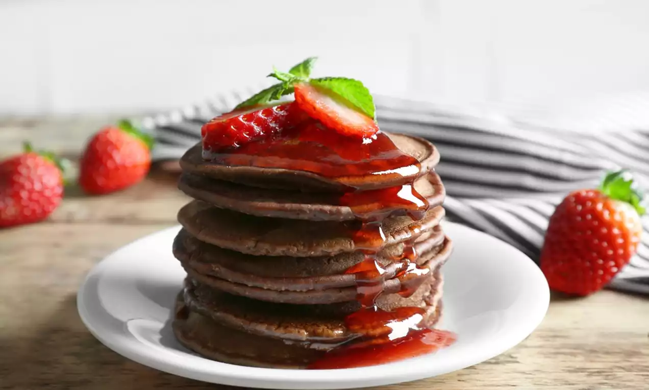 Stack of dark chocolate pancakes layered with glossy red strawberry sauce, topped with fresh strawberry slices and a mint sprig on a white plate