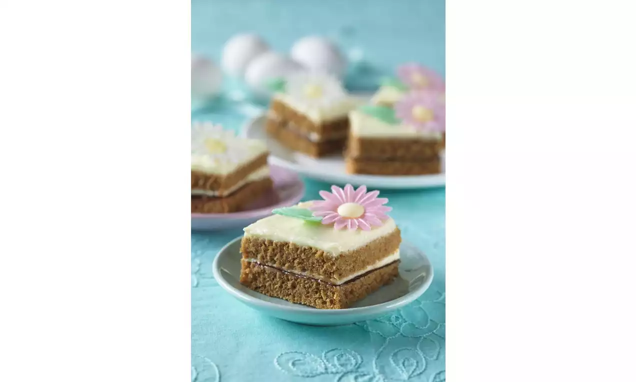 Perníkové rezy s medovo hnedým piškótom, vrstvou bielej polevy a dekoráciou farebných marcipánových kvetov na svetlom tanieri