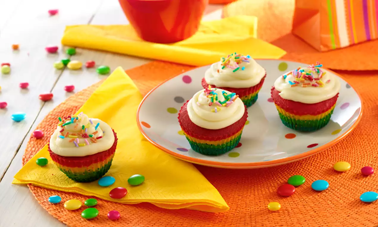 Cupcakes multicolores avec une base rayée arc-en-ciel, crème blanche lisse et éclats de sucre colorés sur une assiette à pois